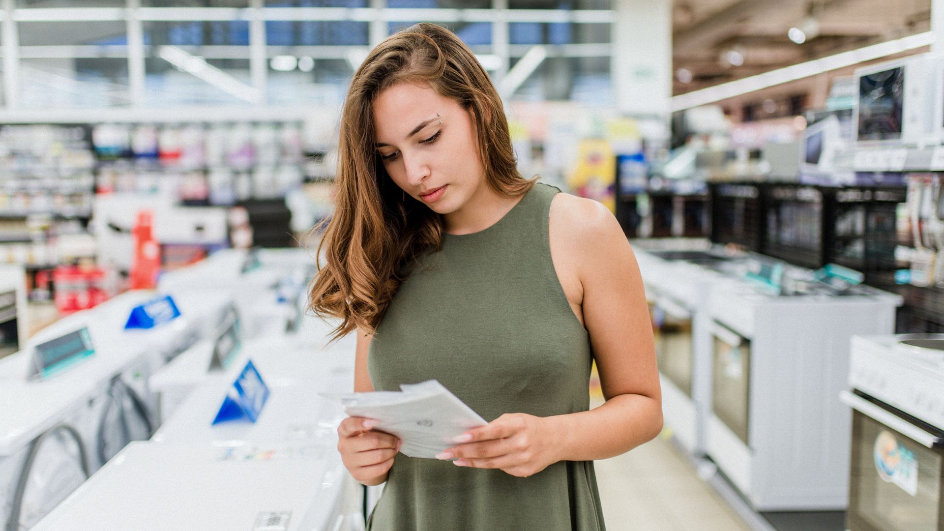 A woman standing in an appliance store, holding and reading a set of papers or brochures